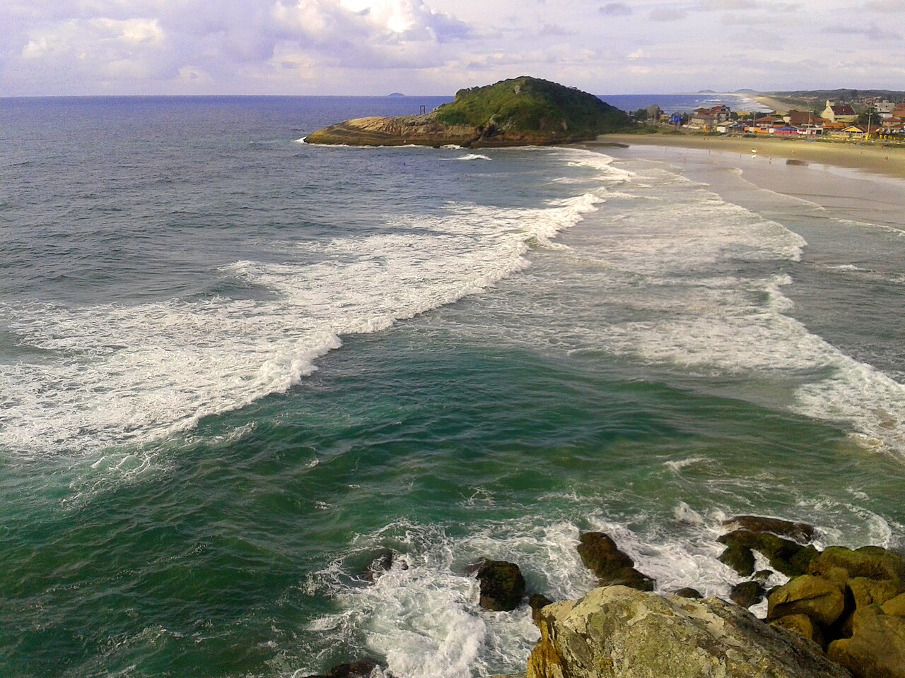 Vista elevada de praia com ondas e espuma, rochas em primeiro plano, promontório vegetado e construções junto à areia.