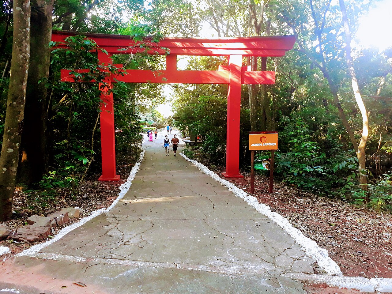 Portal vermelho sobre caminho pavimentado, cercado por vegetação, com visitantes ao fundo e placa indicando Jardim Japonês.