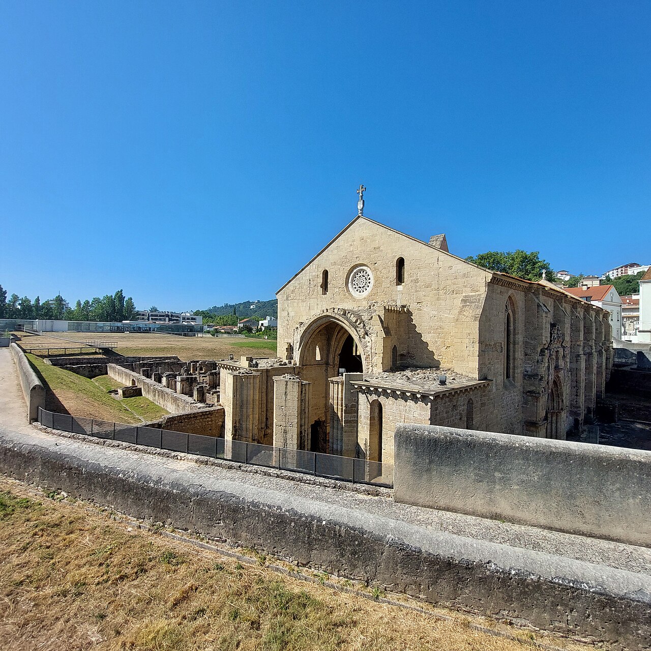 Igreja de Santa Clara-a-Velha com portal ogival e rosácea, cercada por ruínas, bases de colunas, gramados e passarelas.