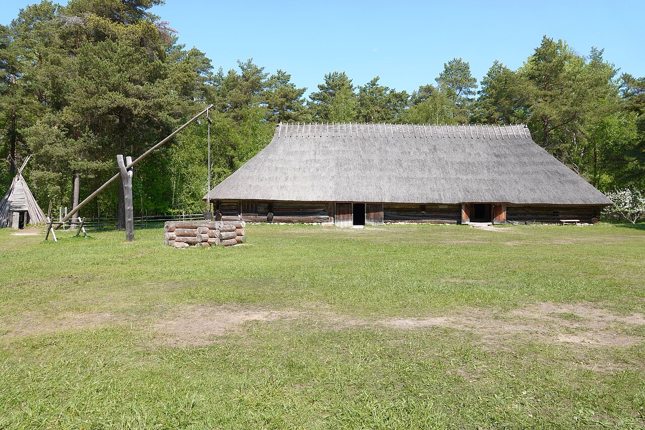 Construção comprida de toras na fazenda Sassi-Jaani, com telhado de palha, portas abertas e gramado diante da fachada; à esquerda, um poço com haste de madeira.