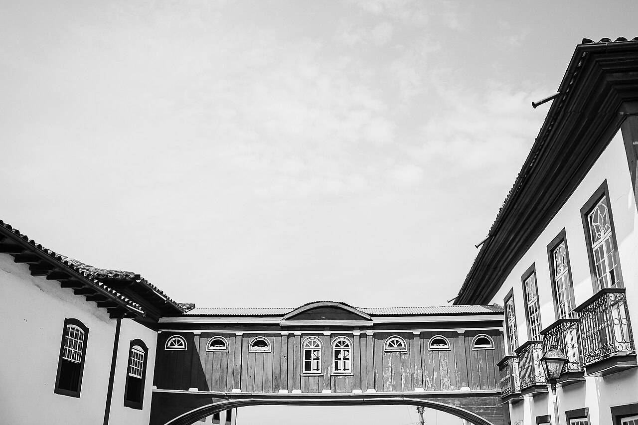 Fotografia em preto e branco de passagem elevada fechada, com janelas, ligando dois sobrados, vista de baixo.