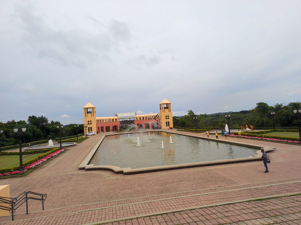 Espelho d’água com chafarizes no Parque Tanguá, cercado por jardins, com construção amarela e alaranjada de duas torres ao fundo.