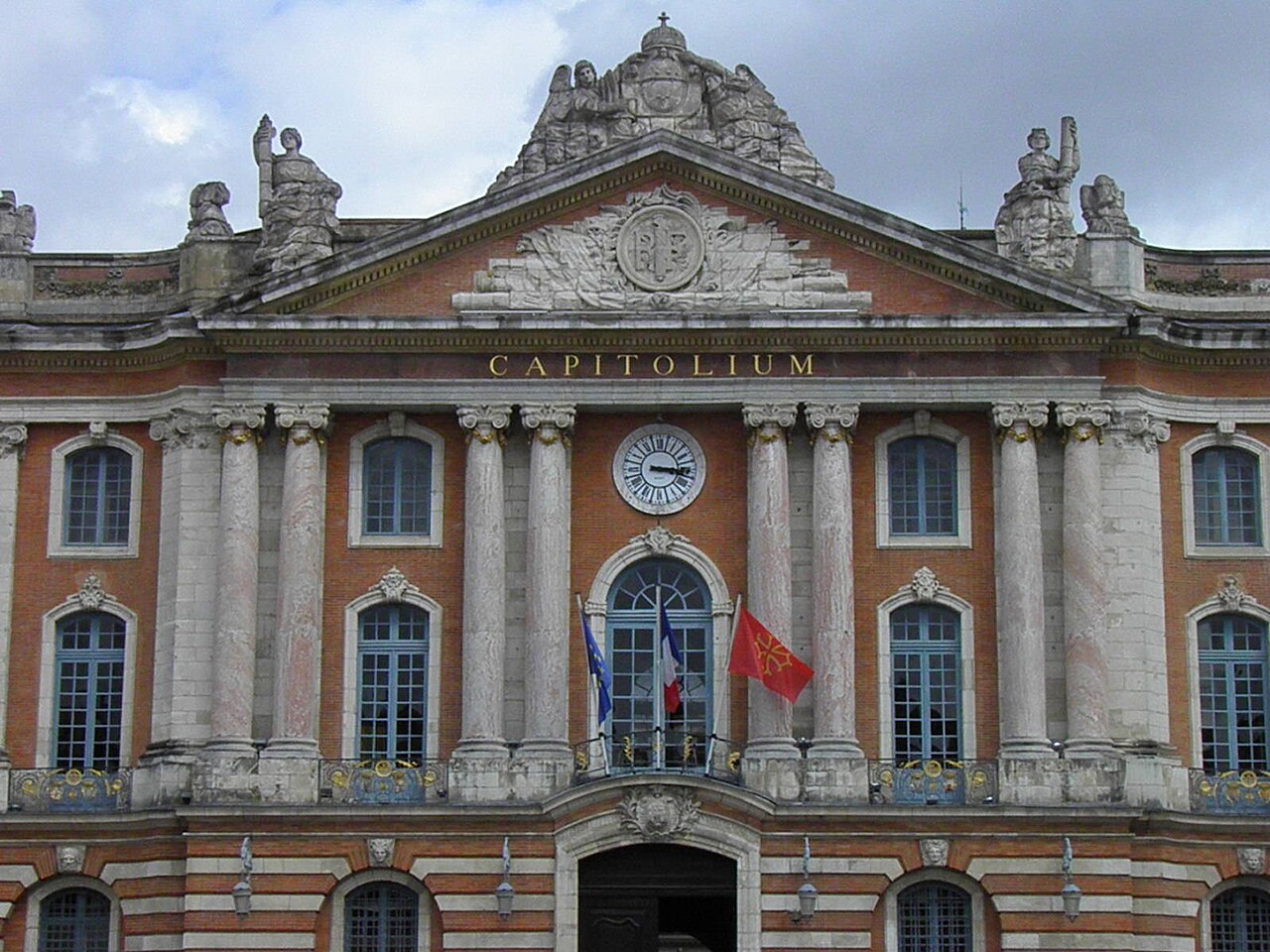 Fachada do Capitole de Toulouse em tijolos e pedra, com colunas, relógio, esculturas e bandeiras.