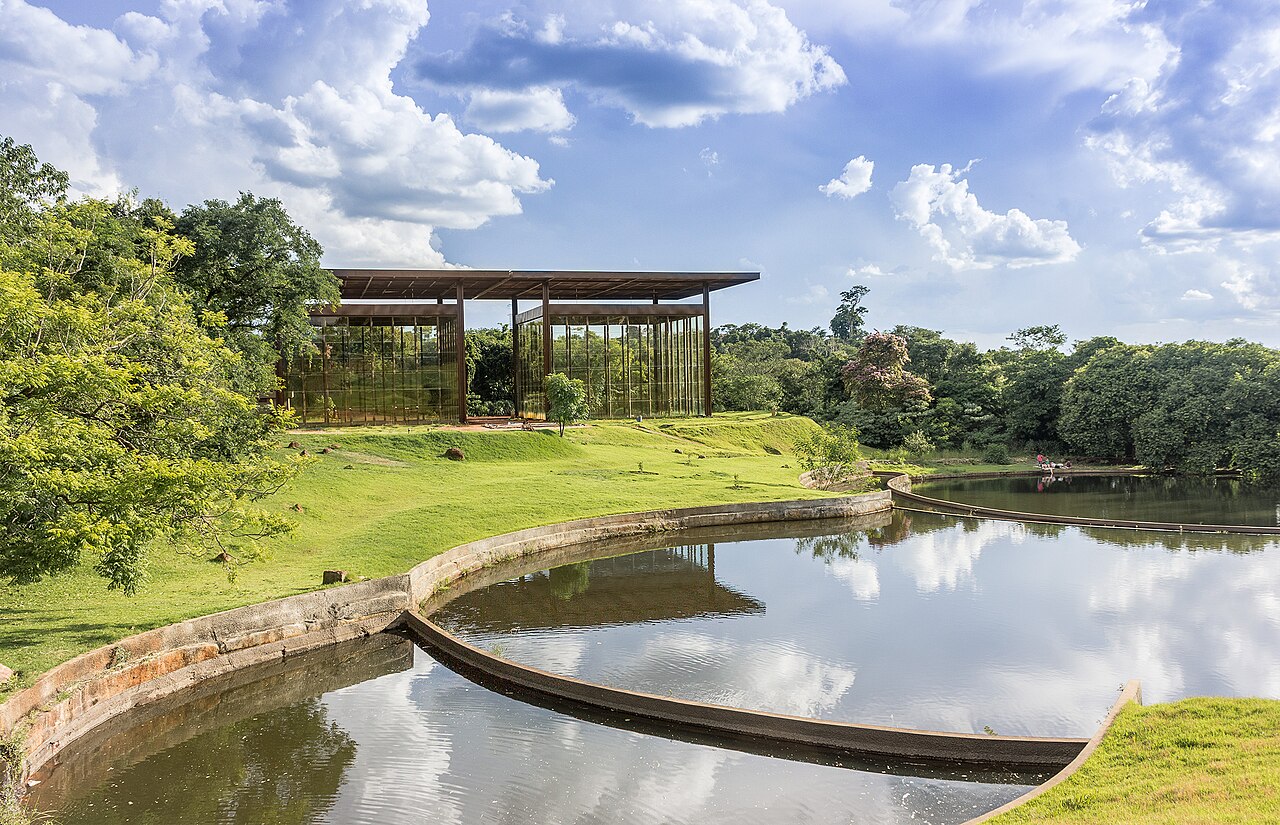 Estrutura envidraçada do Jardim Botânico cercada por gramados e árvores, com reflexos nos espelhos d’água.
