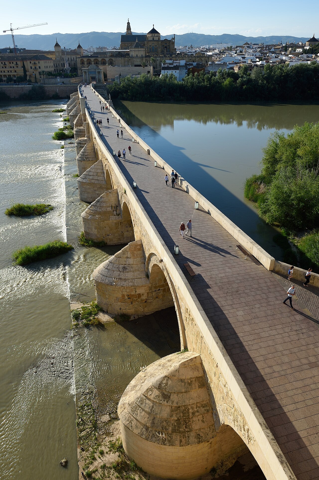 Vista elevada da Ponte Romana sobre o Guadalquivir, com arcos de pedra, pedestres e edifícios históricos de Córdoba ao fundo.