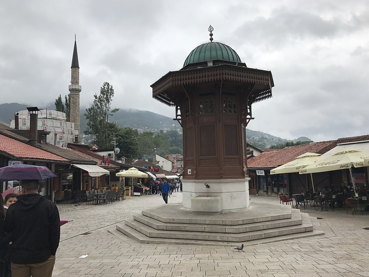 Sebilj com estrutura de madeira, cúpula verde e degraus de pedra, cercado por pessoas e pombos numa praça de Baščaršija.