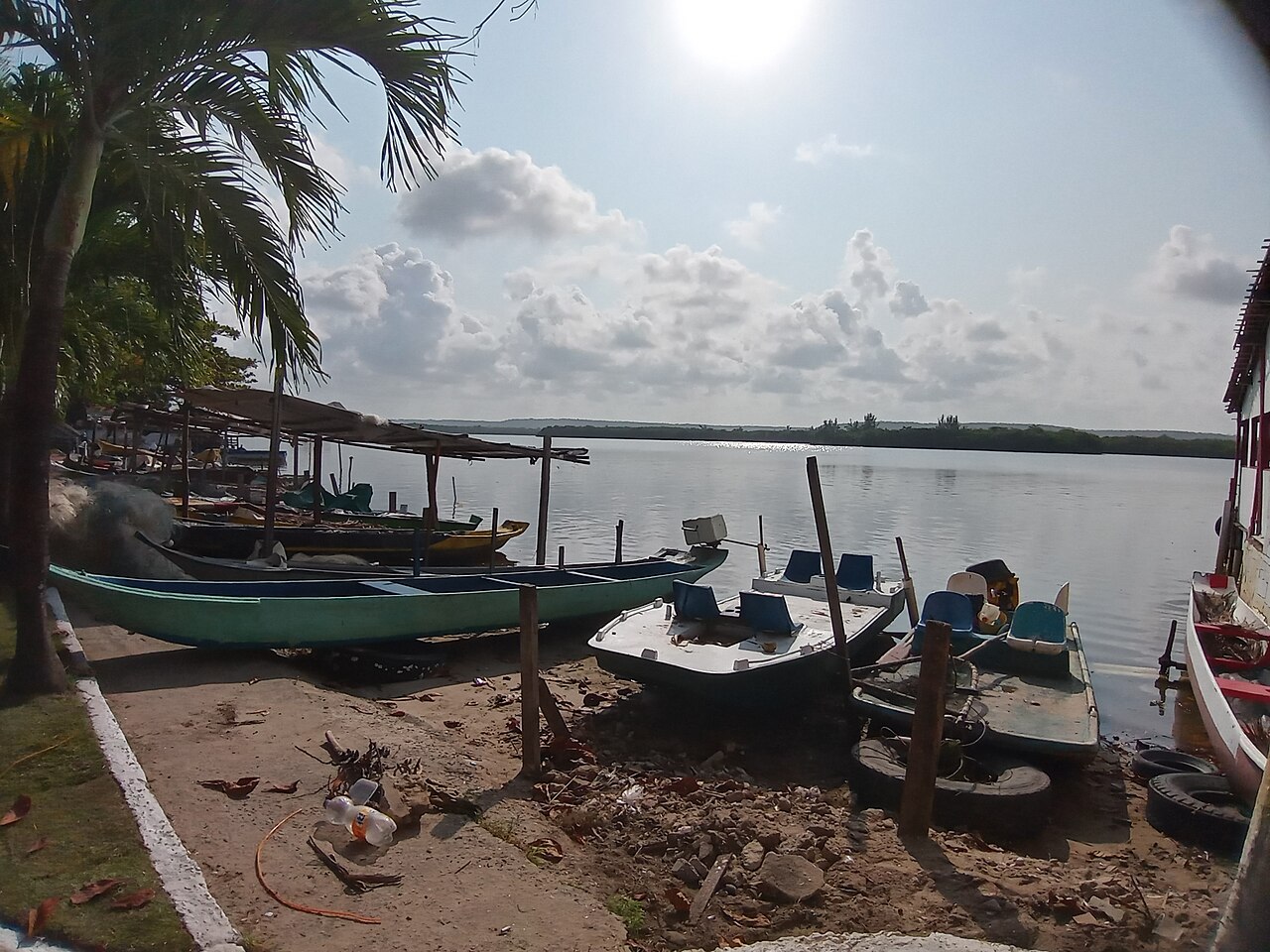 Canoas compridas e pequenas embarcações com assentos azuis junto a uma margem de areia e pedras no Pontal da Barra.