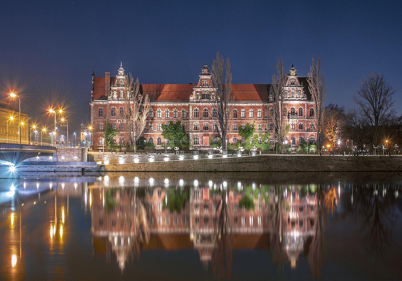 Museu Nacional de Wrocław à noite, com fachada de tijolos vermelhos iluminada, árvores em frente, uma ponte à esquerda e reflexos na água.