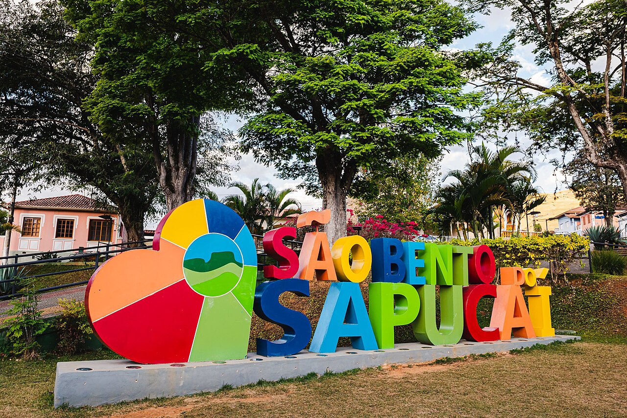 Letreiro multicolorido de São Bento do Sapucaí com um grande coração à esquerda, cercado por gramado, árvores e construções baixas.