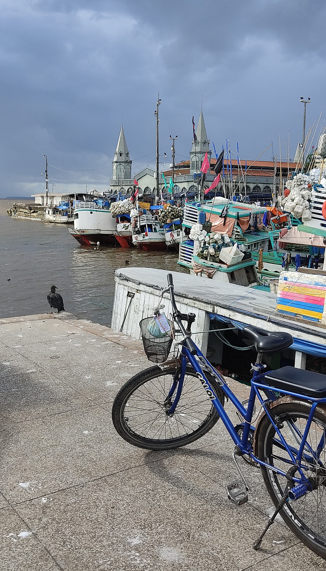 Bicicleta azul no cais, barcos de madeira atracados e edifício com torres pontiagudas ao fundo no Complexo do Ver-o-Peso.
