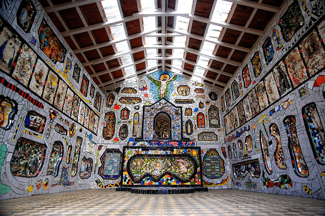 Interior de pequena capela com paredes e altar cobertos por mosaicos coloridos, imagens religiosas e crucifixo acima do altar.