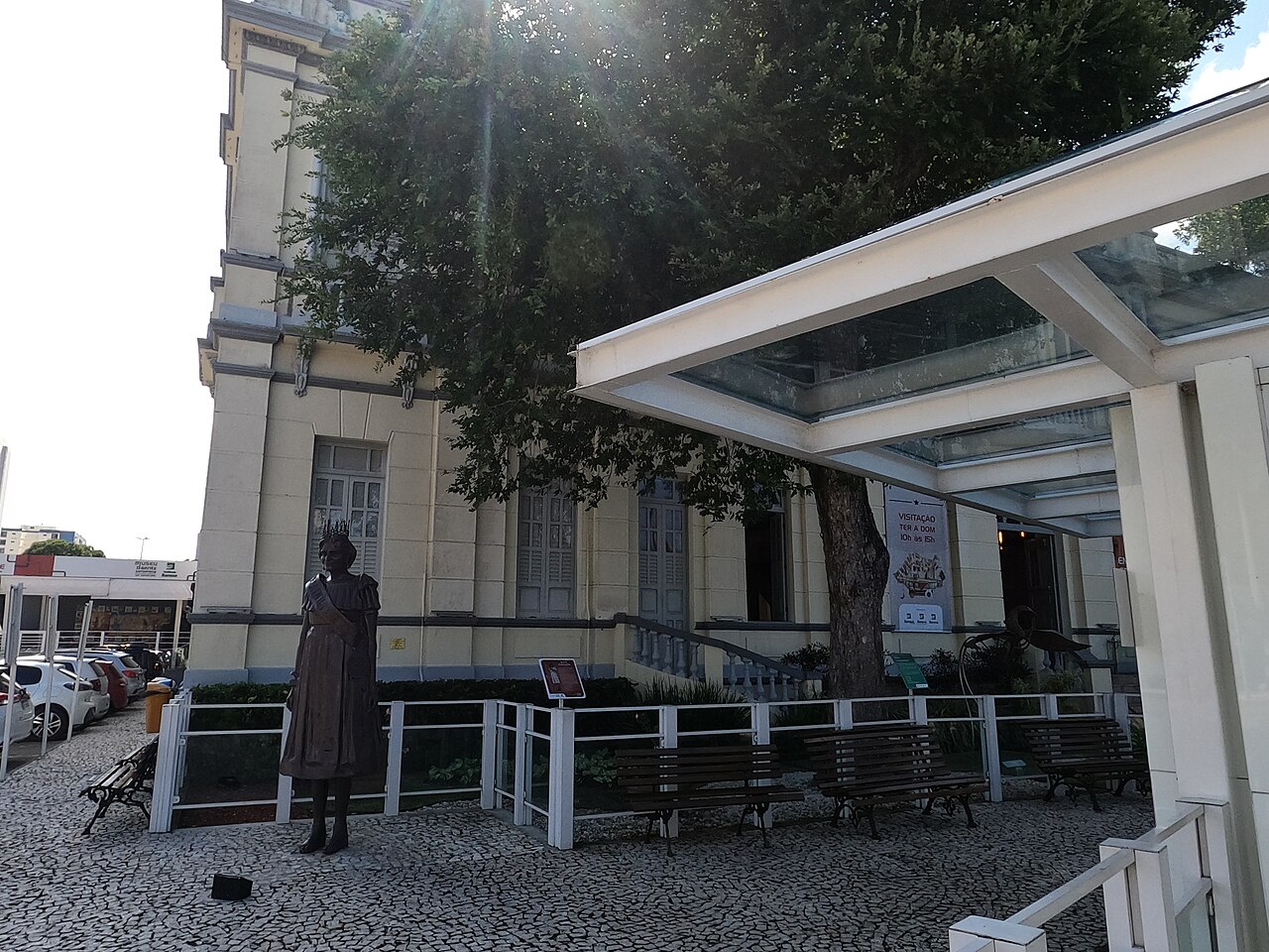 Edifício histórico claro com janelas altas, árvore, escultura de figura feminina, bancos e cobertura de vidro junto à entrada.