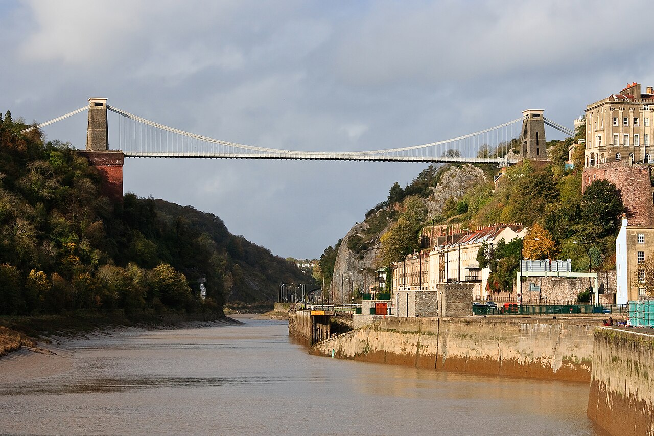 Clifton Suspension Bridge sobre um desfiladeiro, com torres de pedra, encostas arborizadas e um rio com margens de lama expostas.
