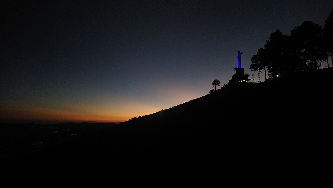 Estátua iluminada em azul sobre uma encosta, à direita, com árvores em silhueta e uma faixa alaranjada no horizonte ao entardecer.