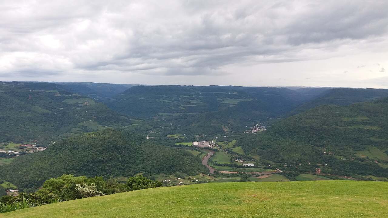 Vista elevada do Ninho das Águias, com gramado, morros verdes, áreas agrícolas, construções dispersas e rio sinuoso sob céu nublado.