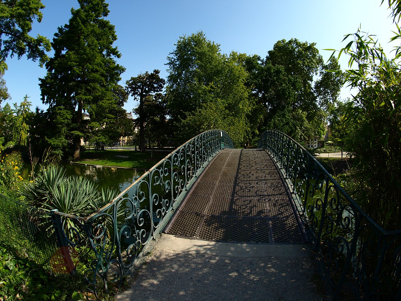 Ponte arqueada com grades ornamentadas sobre um pequeno lago cercado por árvores no Jardin Public.
