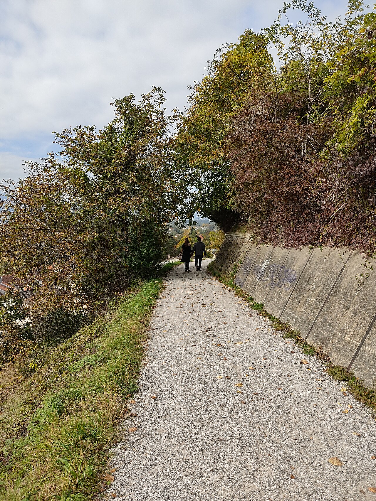 Duas pessoas caminham de costas por um caminho de cascalho em Piramida, entre vegetação e um muro de contenção.