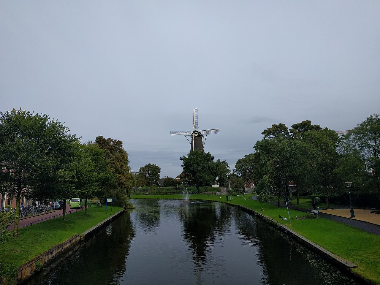 Canal com pequeno chafariz e margens arborizadas, com o moinho de torre De Valk parcialmente visível ao fundo.