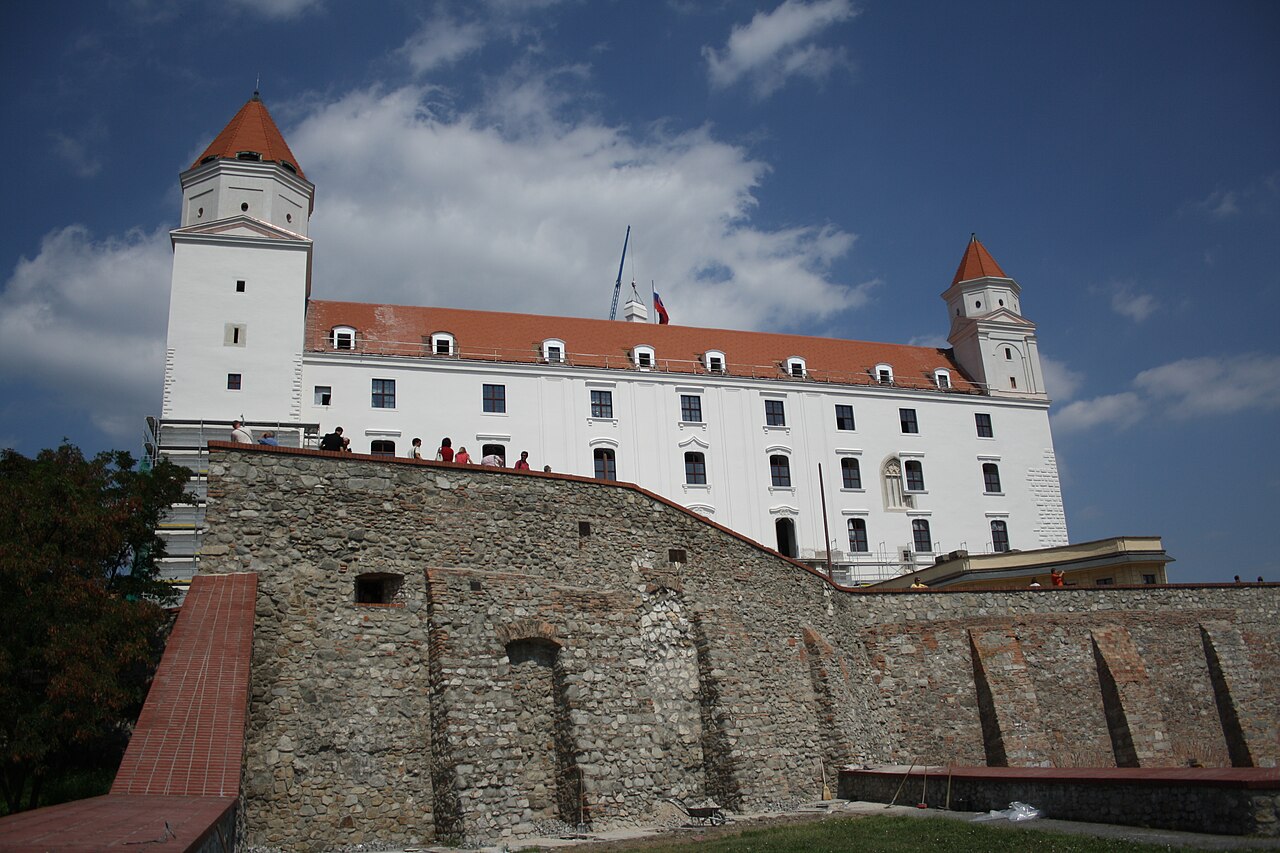 Castelo de Bratislava visto de baixo, com paredes brancas, telhados avermelhados, muralhas, visitantes e estruturas de obra.