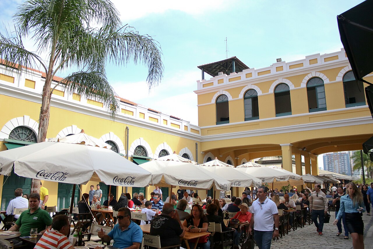 Pátio interno do Mercado Público de Florianópolis com mesas, guarda-sóis, palmeiras e fachadas amarelas com portas verdes.