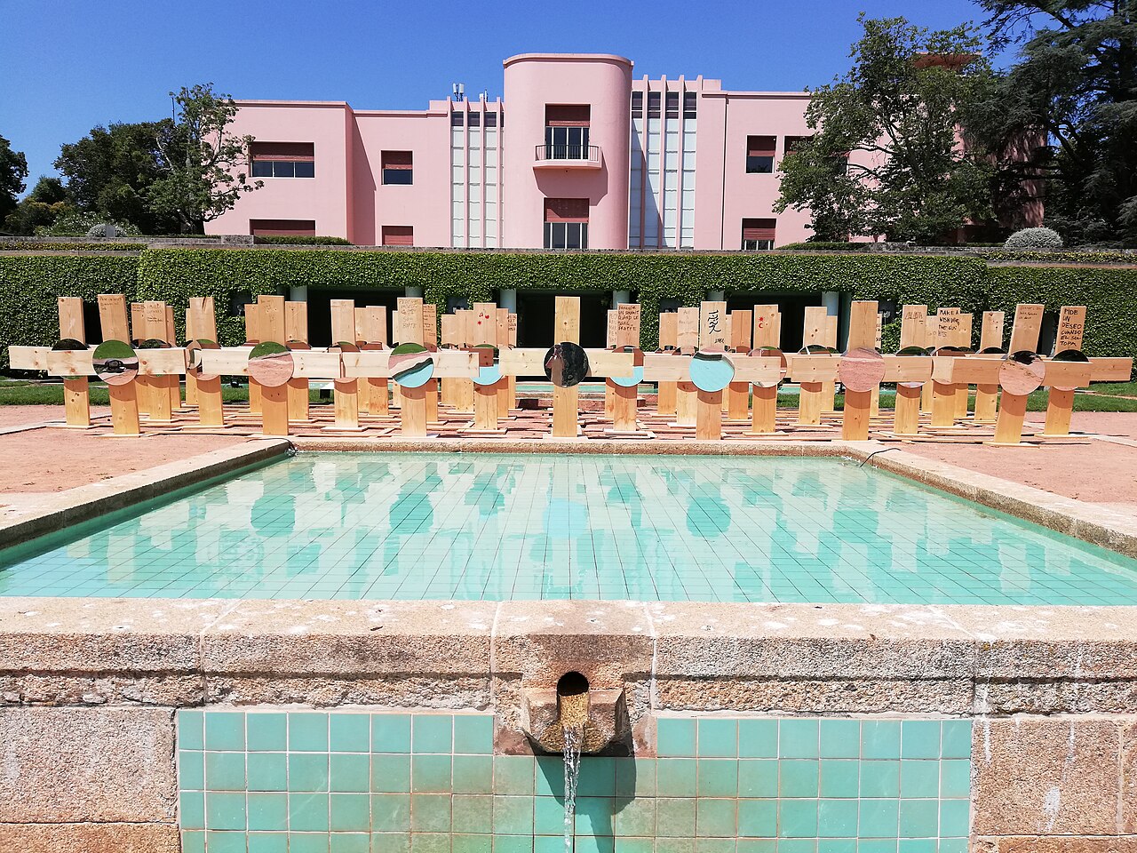 Casa de Serralves rosa atrás de uma sebe, com espelho d’água de azulejos turquesa e uma instalação de madeira em primeiro plano.