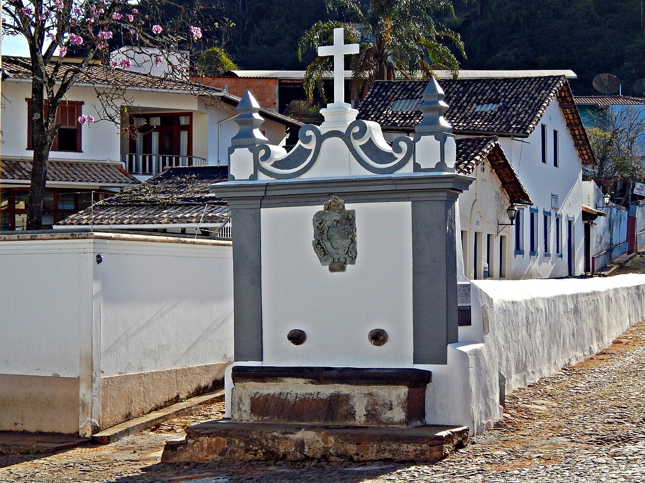 Chafariz branco com molduras cinza, duas bicas, tanque, brasão central e cruz, cercado por calçamento e casas.