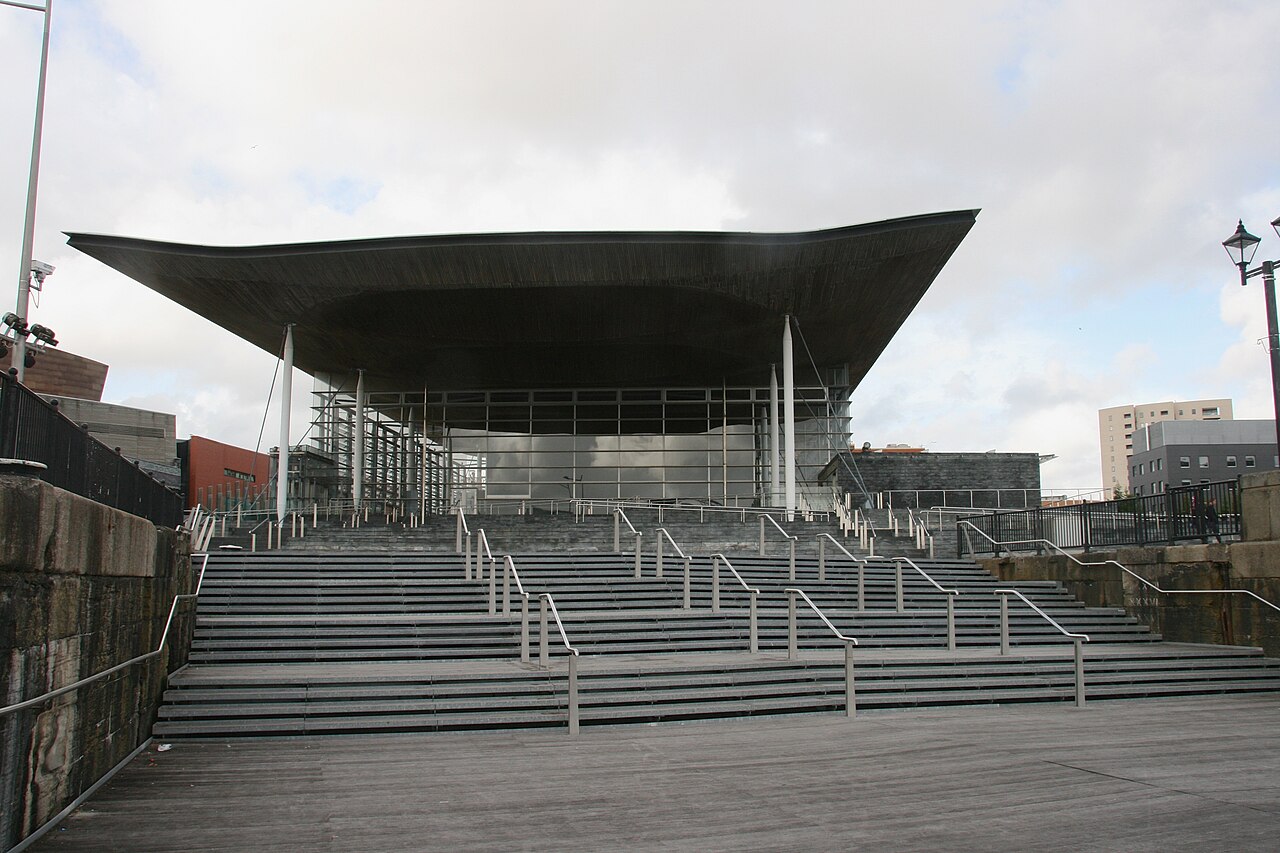 Fachada envidraçada do Senedd, com colunas esbeltas, ampla cobertura de madeira e escadaria com corrimãos metálicos.