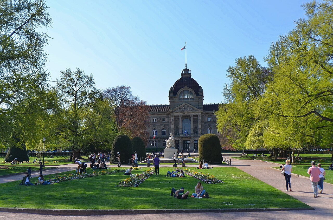 Jardim da Place de la République com caminhos, flores, pessoas e monumento diante da fachada simétrica do Palais du Rhin.