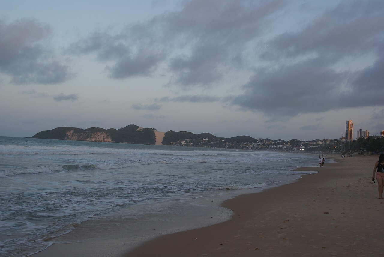Praia com ondas, areia úmida e pessoas caminhando, com edifícios à direita e o Morro do Careca ao fundo sob céu nublado.