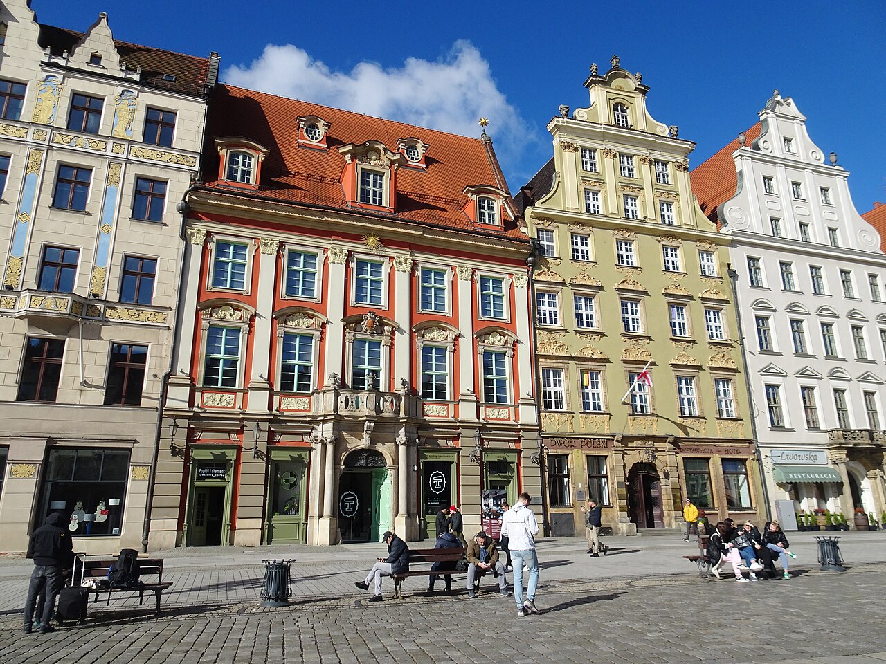 Praça pavimentada do lado oeste do Rynek, com bancos e pedestres diante de fachadas ornamentadas em vermelho, bege e branco, sob céu azul com nuvens.