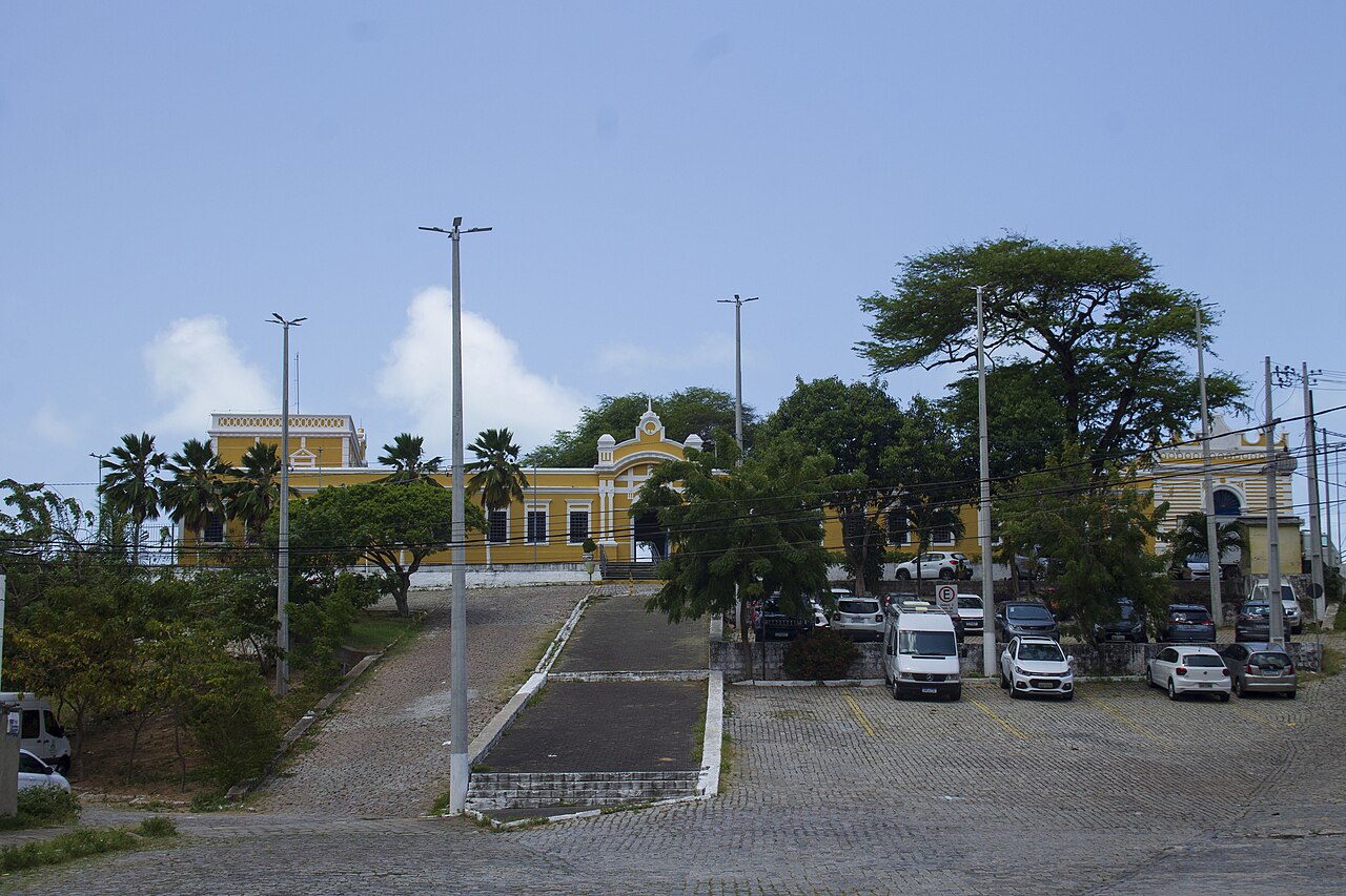 Edifício histórico amarelo com detalhes brancos e janelas altas, parcialmente encoberto por árvores e palmeiras, com acessos pavimentados e veículos à frente.