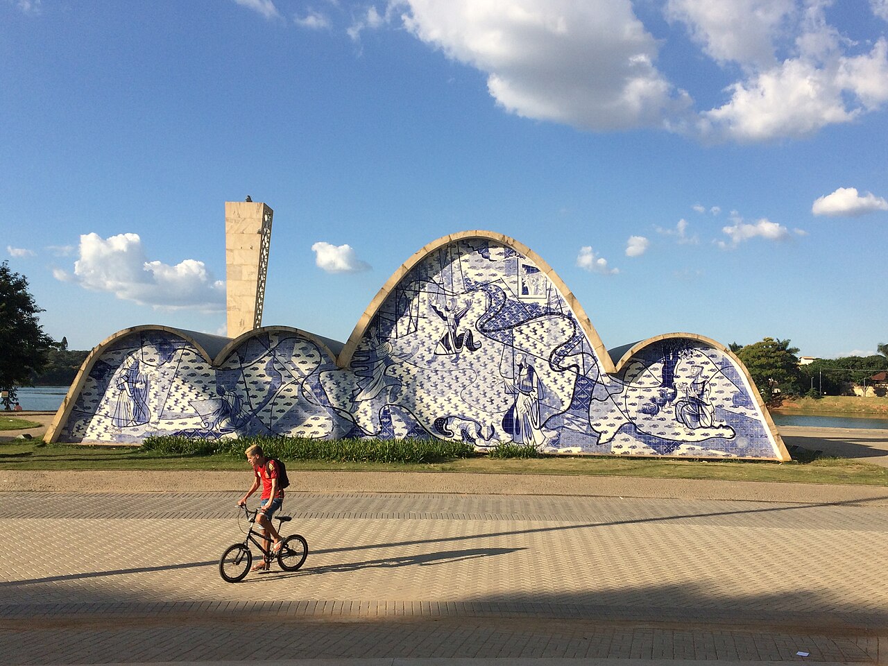 Exterior da Igreja São Francisco de Assis com volumes curvos, painel de azulejos azuis e brancos, gramado e ciclista no passeio.