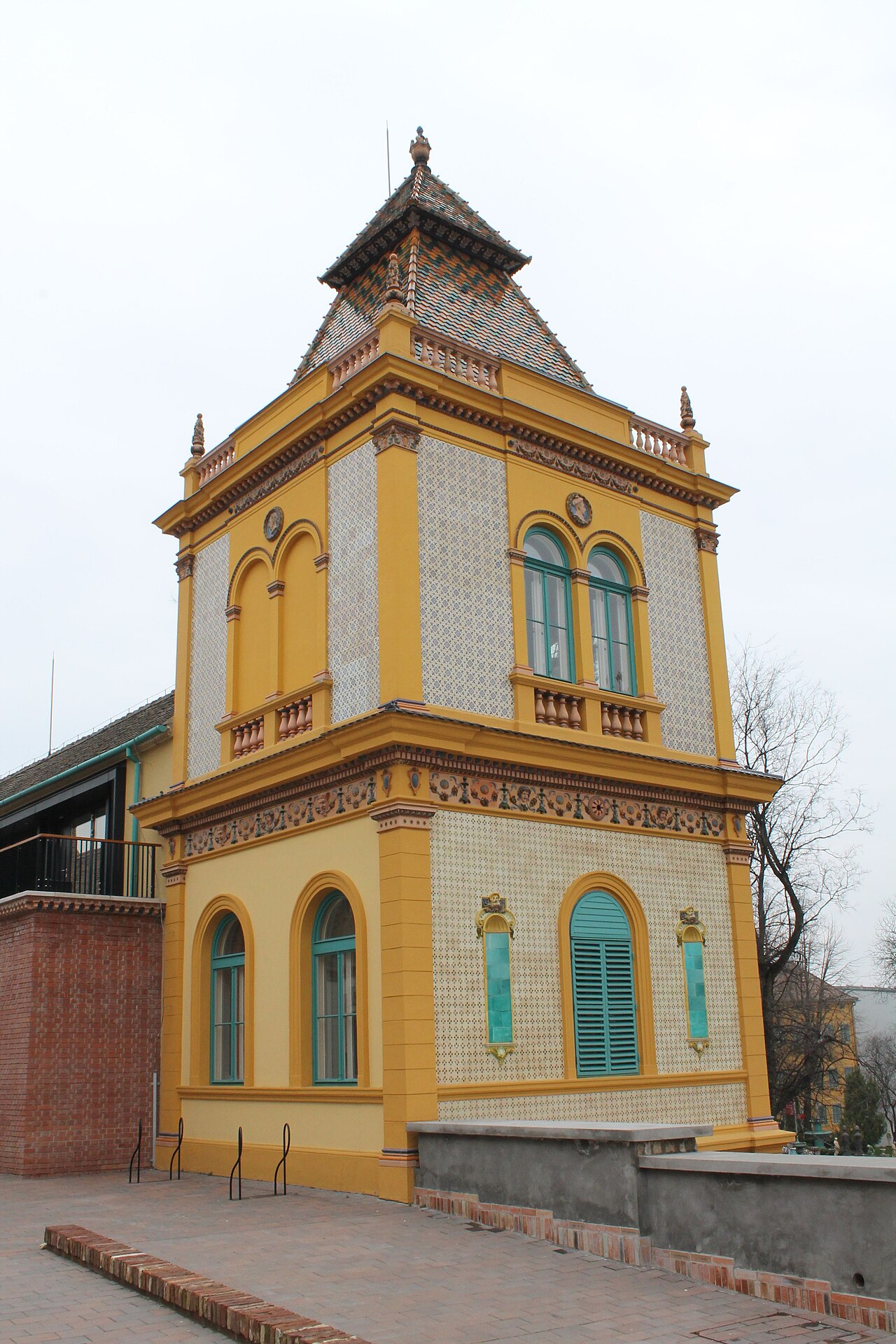 Edifício amarelo do conjunto Zsolnay com painéis de azulejos, janelas verde-turquesa e torre de telhas multicoloridas.
