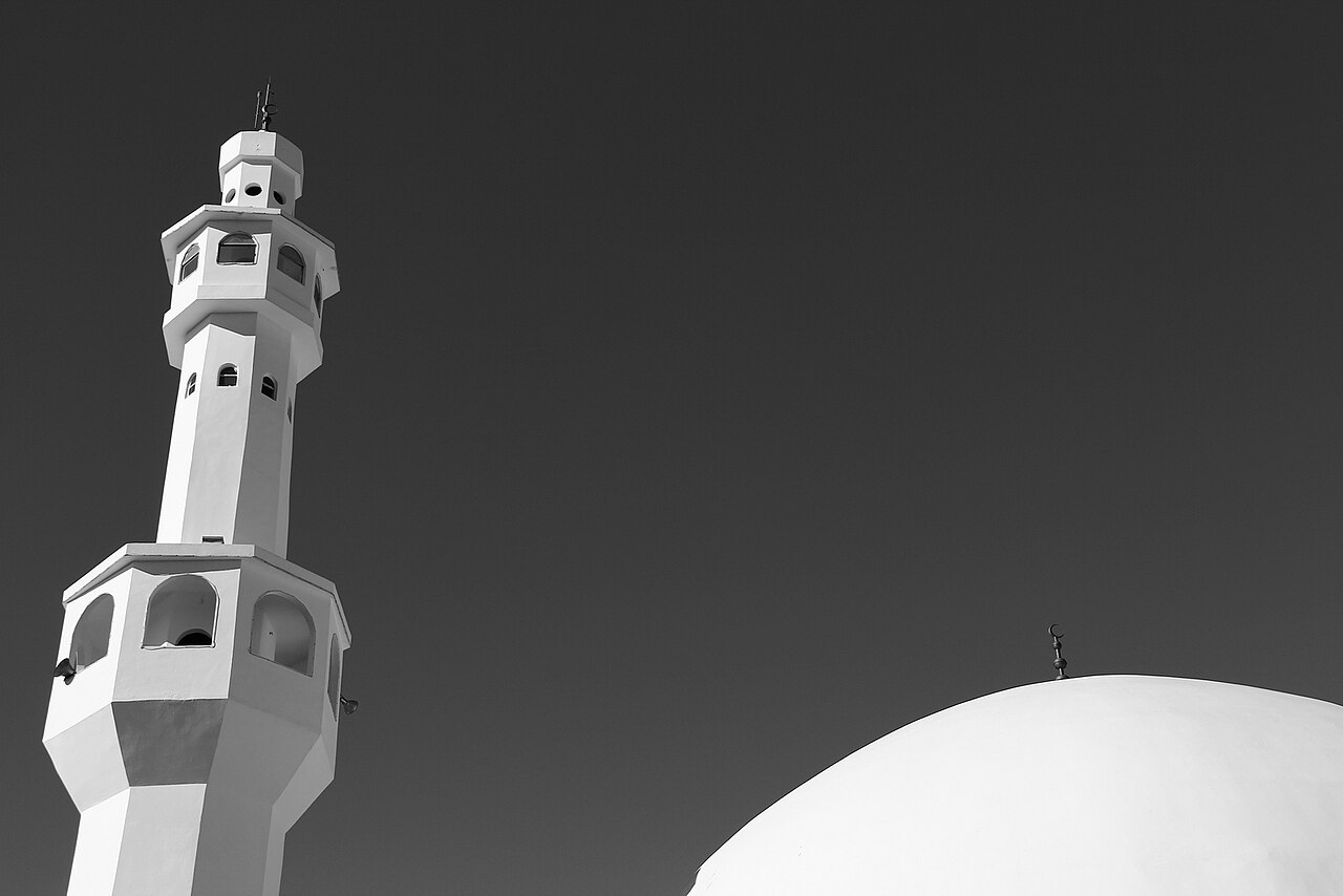 Fotografia em preto e branco de um minarete à esquerda e parte de uma cúpula com crescente à direita, vistos de baixo.