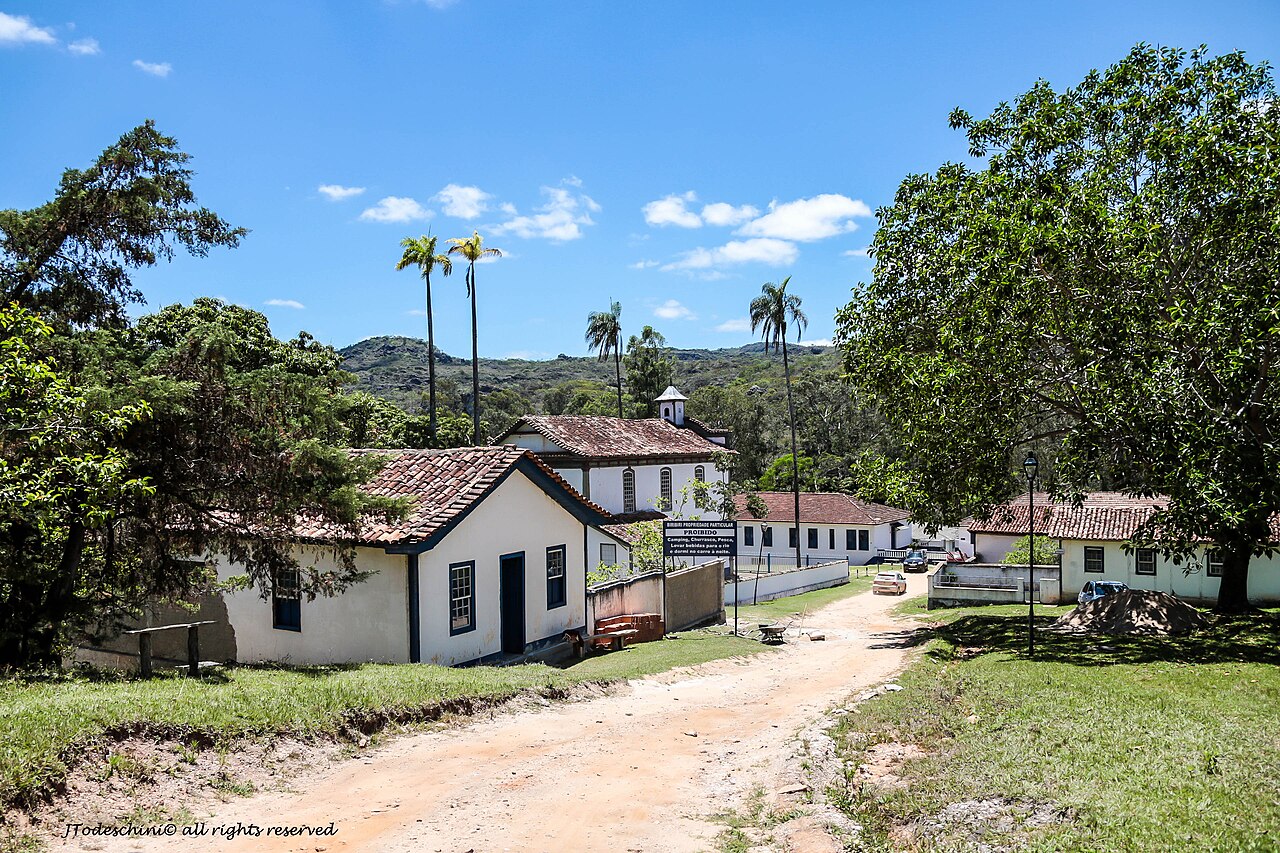 Casas brancas com telhados cerâmicos, igreja ao fundo, rua de terra, palmeiras e morros vegetados na Vila de Biribiri.