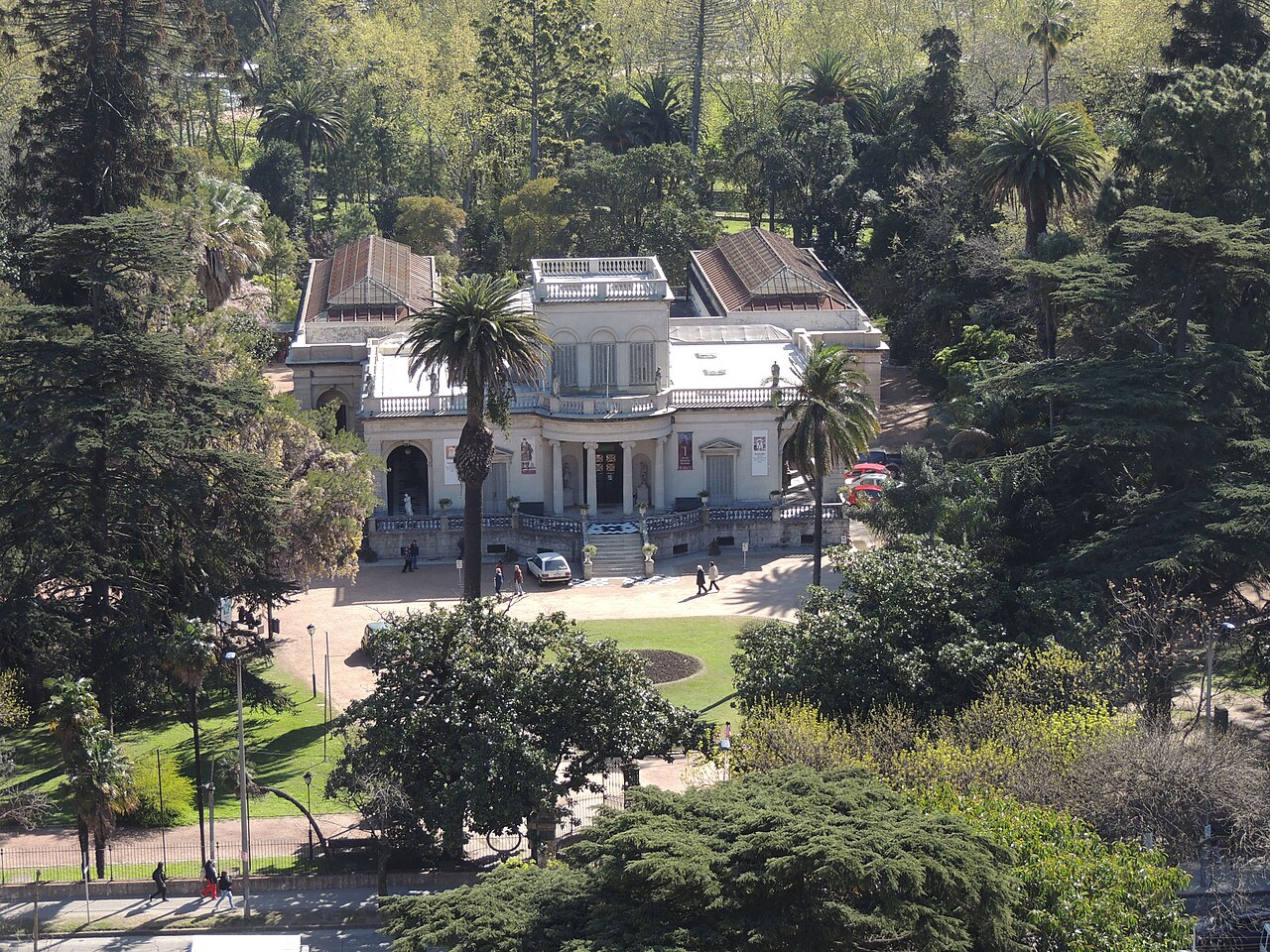 Casarão claro do Museo Blanes visto de cima, com fachada simétrica, colunas, escadaria central, palmeiras e jardins ao redor.