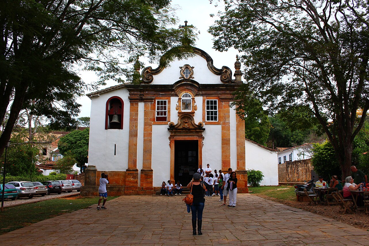 Fachada branca da Igreja de Nossa Senhora do Rosário, com detalhes em ocre, cruz no frontão e pessoas no adro de pedra.