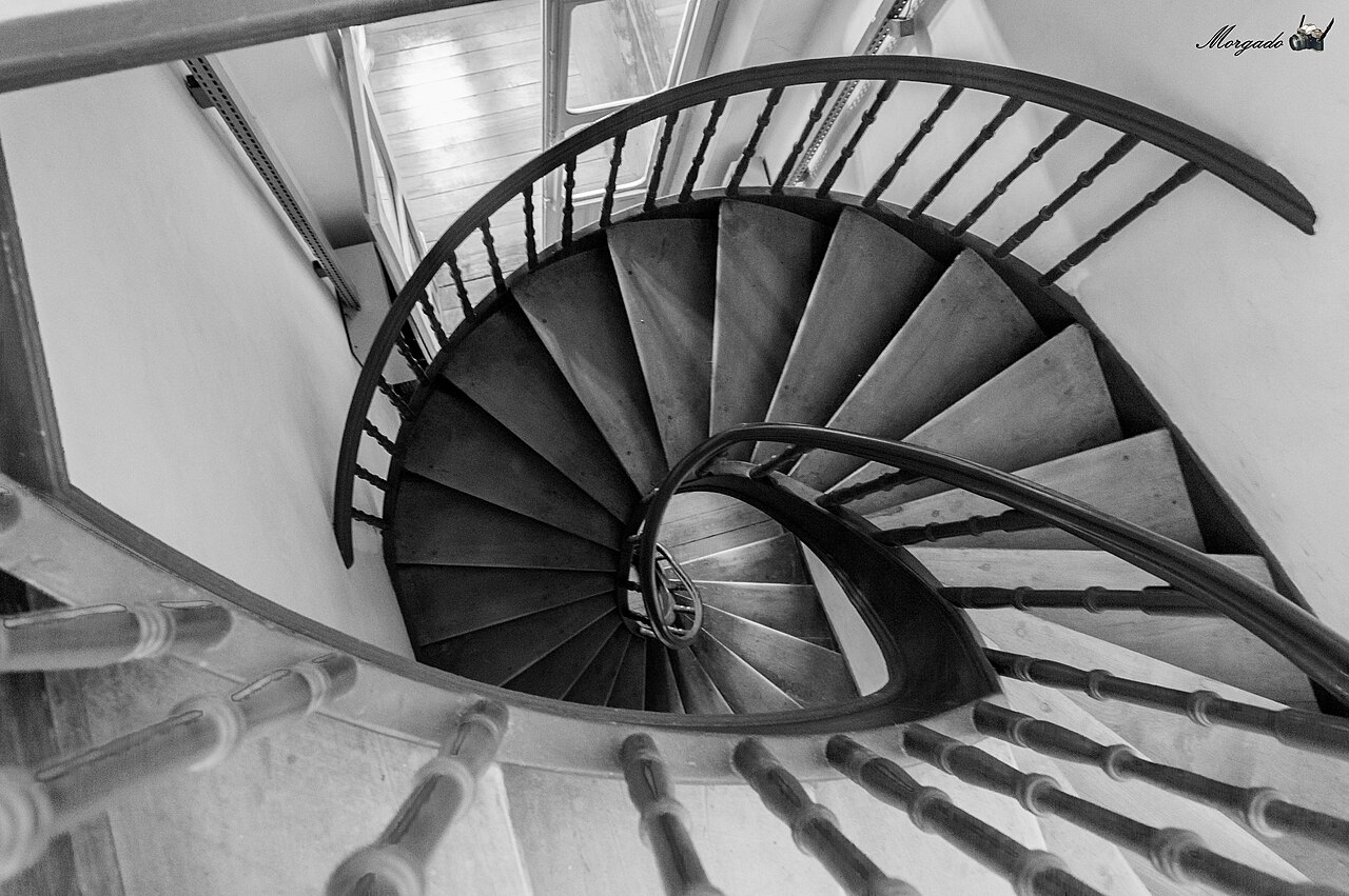Fotografia em preto e branco de escada de madeira em espiral vista de cima, com corrimãos curvos e balaústres torneados, identificada no acervo como interior do Museu do Doce.