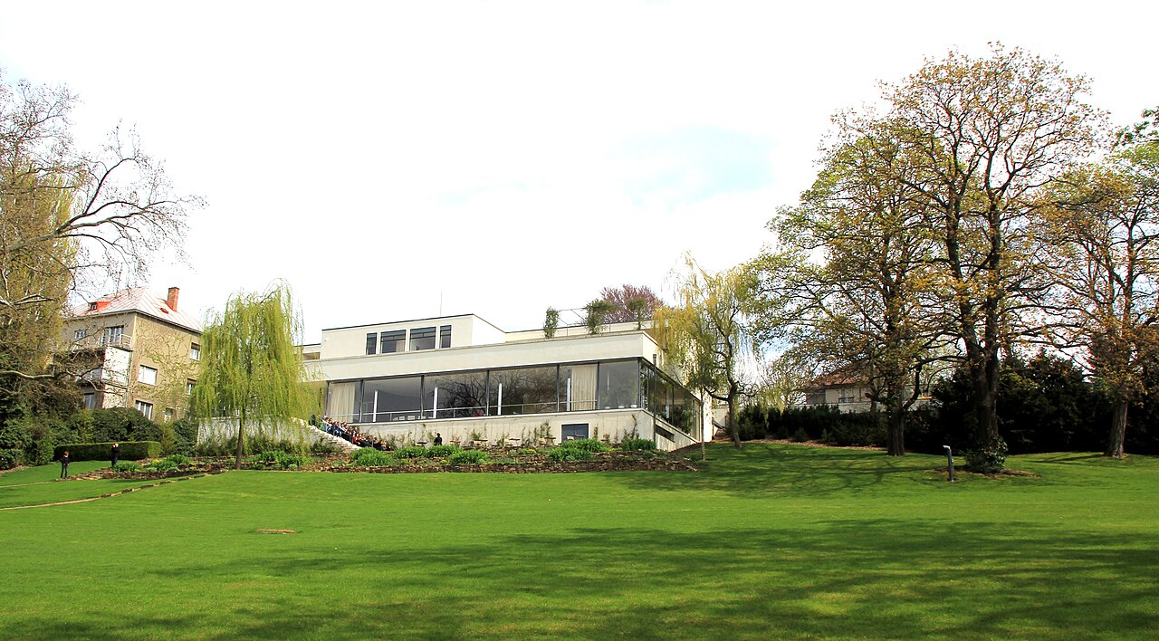 Villa Tugendhat branca, de cobertura plana e fachada envidraçada, diante de um gramado inclinado com árvores e visitantes.