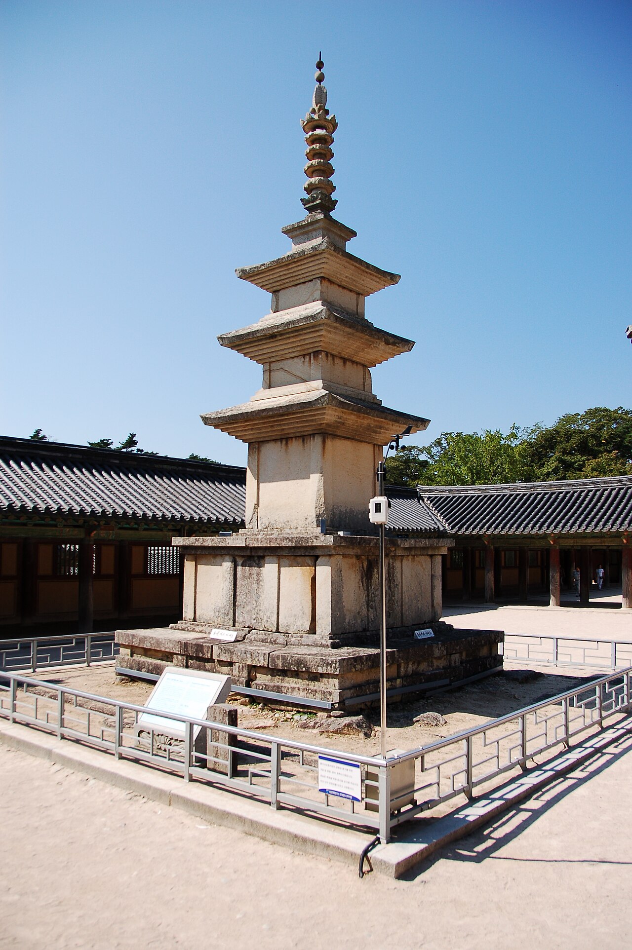 Pagode de pedra Seokgatap, com três andares e remate ornamentado, cercado por grades, com telhados e árvores ao fundo.