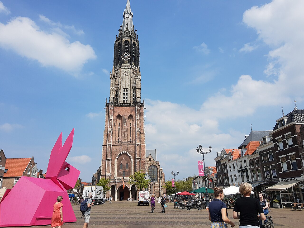 Torre da Nieuwe Kerk diante da praça Markt, com escultura rosa de coelho à esquerda, pedestres e mesas de cafés à direita.