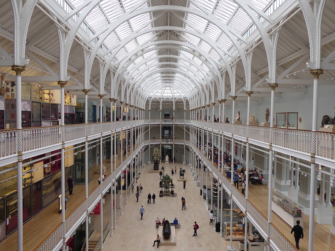 Átrio do National Museum of Scotland com colunas e arcos brancos, cobertura envidraçada, galerias em vários níveis e visitantes.