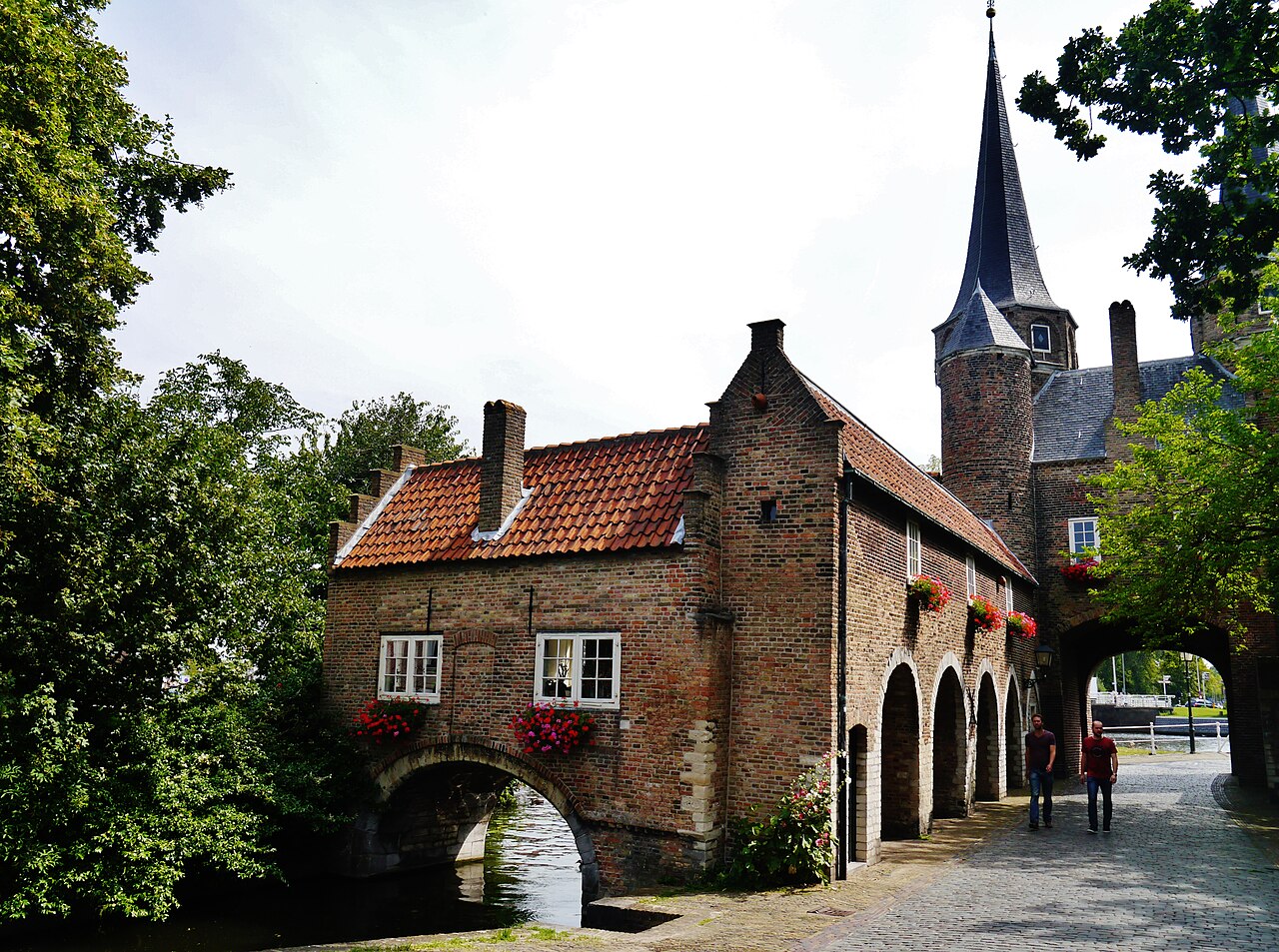 Exterior de tijolos da Oostpoort junto a um canal, com telhados vermelhos, torre pontiaguda, passagem em arco e galeria de arcadas.