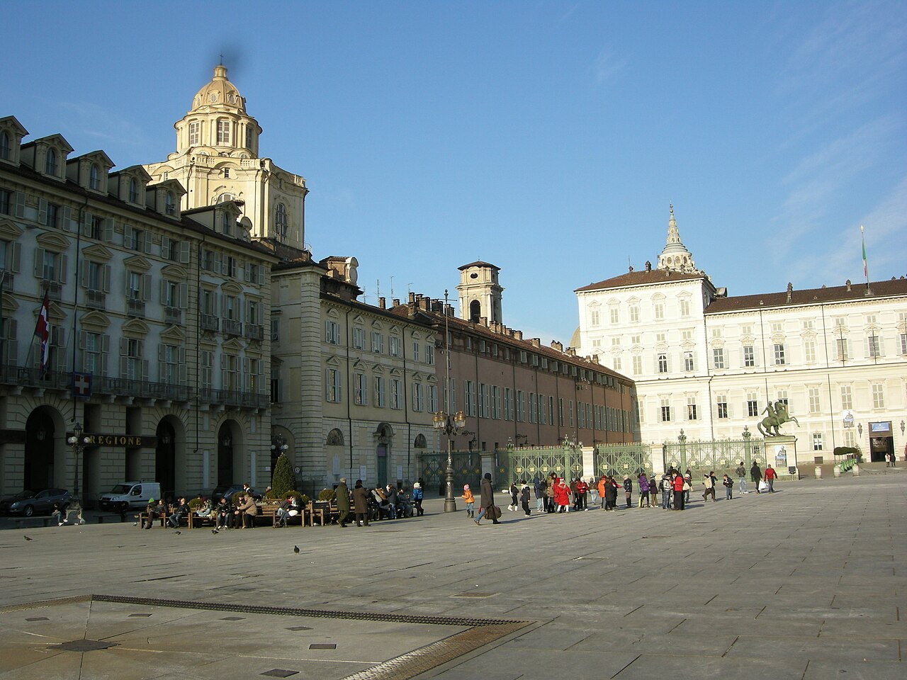 Praça ampla em Piazza Castello, com pedestres, fachadas históricas, cúpula à esquerda e palácio claro com gradil à direita.