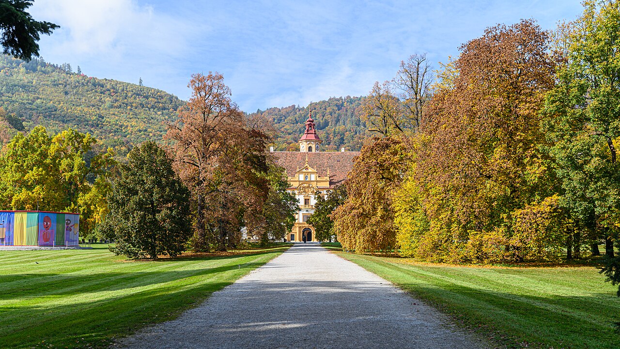 Alameda de cascalho entre gramados e árvores verdes, amarelas e castanhas conduz ao Palácio Eggenberg, parcialmente encoberto pela vegetação.