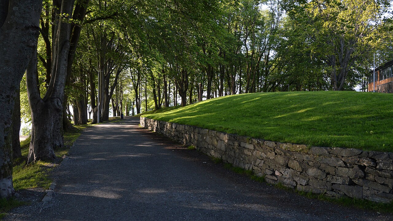 Caminho pavimentado sob árvores altas em Nordnesparken, com muro baixo de pedras e gramado elevado à direita.
