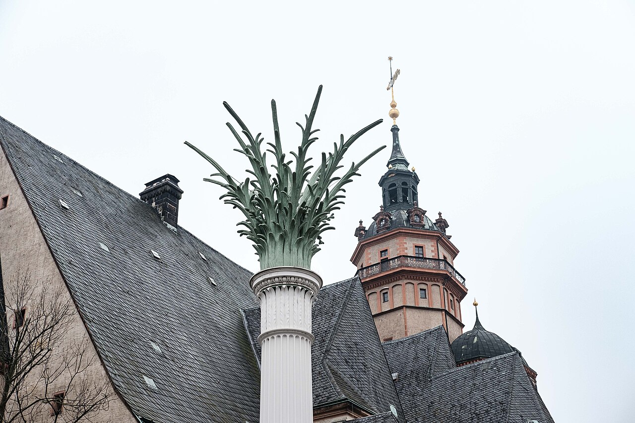 Coluna branca com coroamento verde em forma de palmeira diante dos telhados e da torre da Nikolaikirche.