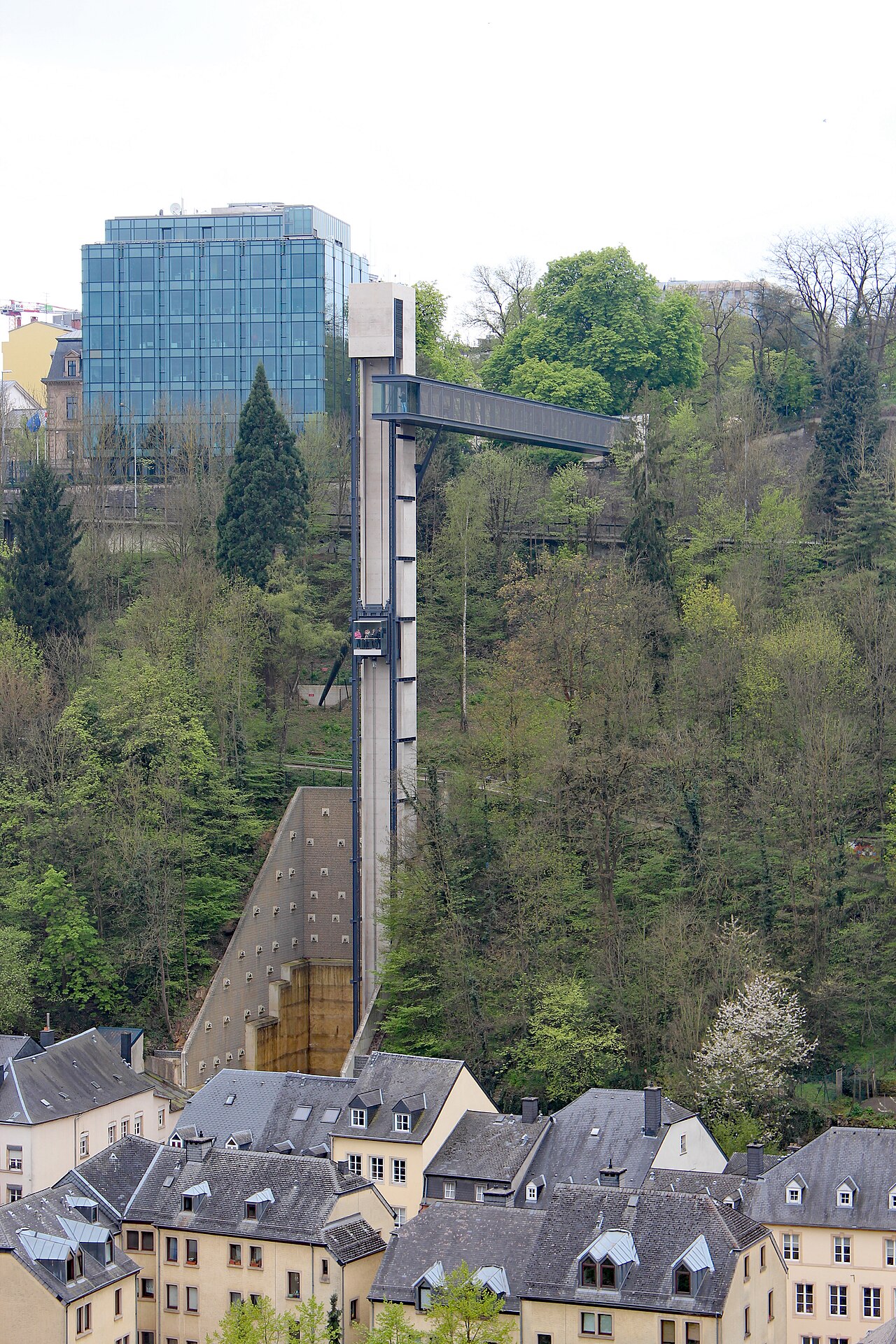 Torre do elevador panorâmico de Pfaffenthal com cabine envidraçada e passarela elevada sobre uma encosta arborizada, acima de casas claras.