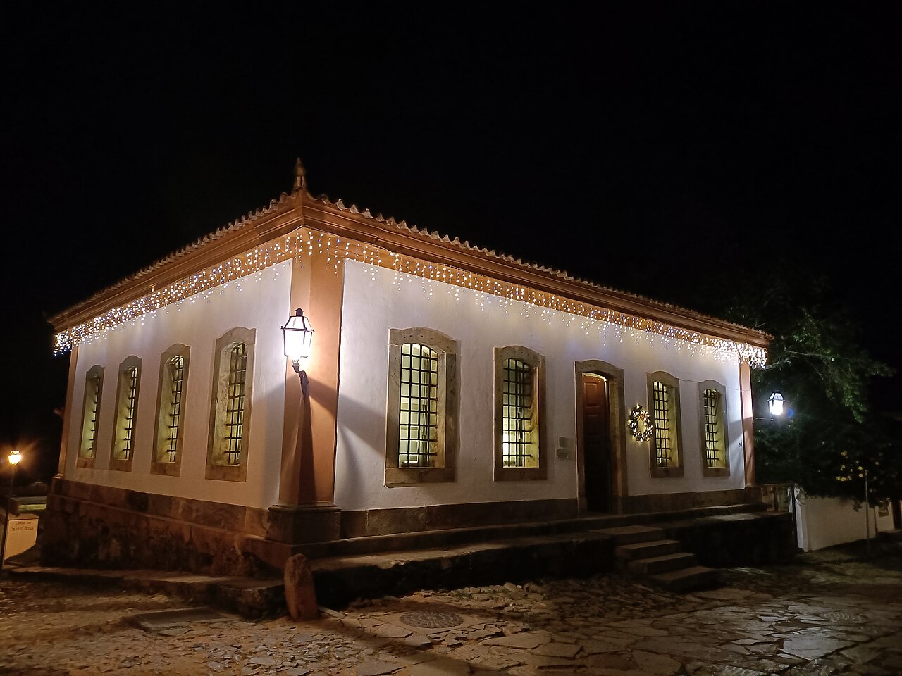 Fachada noturna do Museu de Sant’Ana, em edifício branco de esquina com janelas gradeadas, porta de madeira e luzes nos beirais.