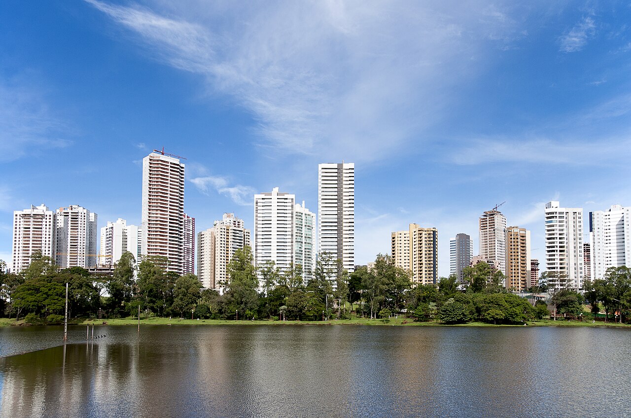 Lago Igapó em primeiro plano, com margem arborizada, gramado e edifícios altos ao fundo sob céu azul.